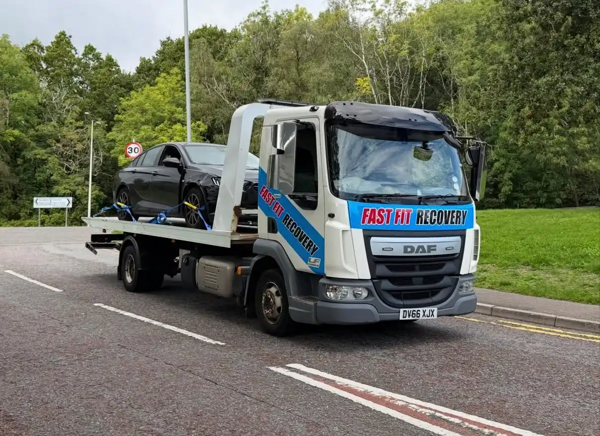 Fast Fit Recovery DAF breakdown truck carrying a black car strapped down on its bed, driving on a tree-lined road