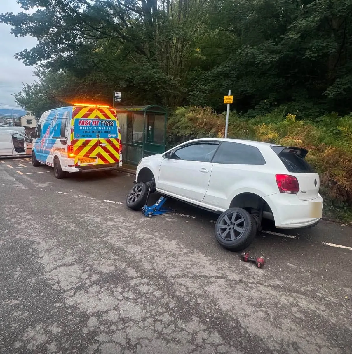 Fast Fit Tyres van with its amber beacons and rear chevrons on at the roadside while a white hatchback with a front wheel off sits on a jack