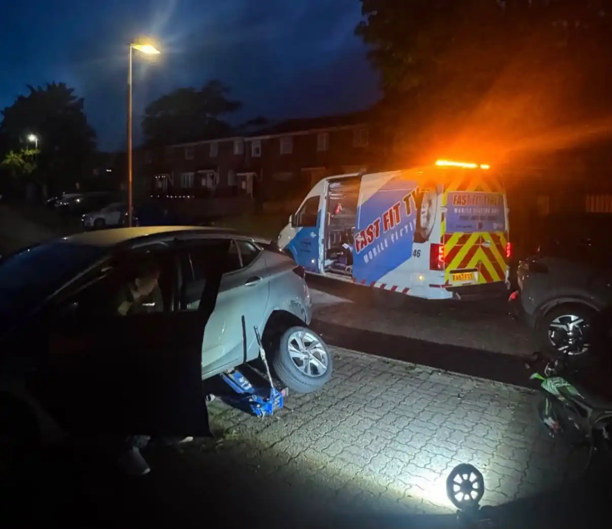 Fast Fit mobile tyre fitting van with its beacons on, parked behind a car jacked up for a new tyre on a residential street at night