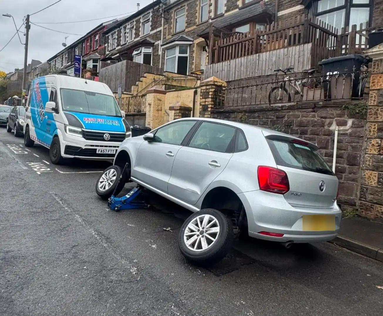 Fast Fit Tyres van parked on a terraced street behind a silver hatchback jacked up with two wheels off for new tyres